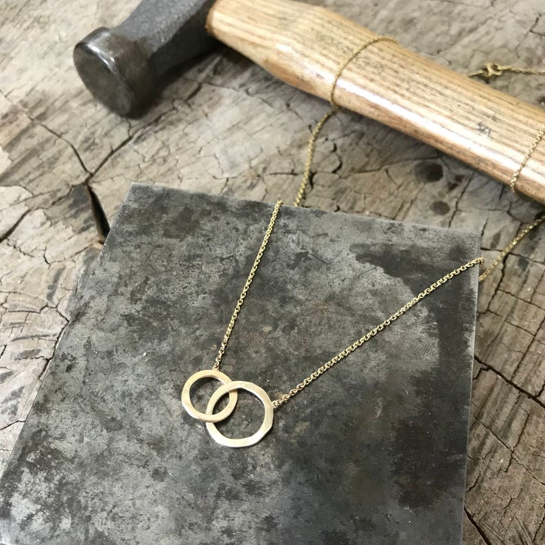 May include: A gold necklace with a delicate chain and a pendant featuring two interlocking circles. The necklace is displayed on a gray surface with a wooden background.