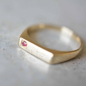 May include: A gold band ring with a small red gemstone set in the center.