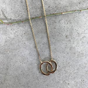 May include: A gold chain necklace with a double circle pendant. The circles are intertwined and have a hammered texture.