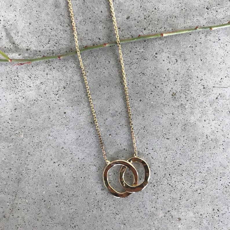 May include: A gold chain necklace with a double circle pendant. The circles are intertwined and have a hammered texture.