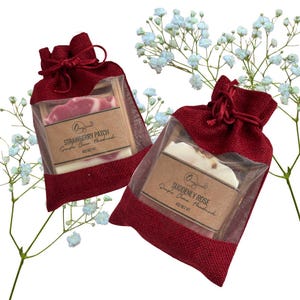 May include: Two handmade soaps in red burlap bags with drawstring closures. One soap is labeled "Strawberry Patch" and the other "Suddenly Rose." Both are in clear plastic sleeves. White flowers are in the background.