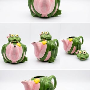May include: A ceramic teapot shaped like a green frog with a pink belly, yellow flower detail, and a pink bow on its head. The teapot lid is the frog's head, which can be removed to reveal the interior. The teapot features a spout and handle.