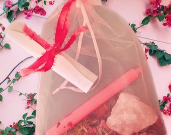 May include: A pink organza bag filled with a pink candle, a pink crystal, dried herbs, and a rolled up piece of paper. The bag is tied with a red ribbon.