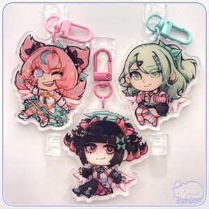 May include: Three acrylic charm keychains featuring anime-style characters with pink, green, and black hair. Each charm has a unique design and is attached to a colored clasp. The charms are displayed against a white background.