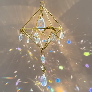 May include: A gold geometric crystal suncatcher with iridescent crystals hanging from a chain. The suncatcher casts rainbows on the wall.