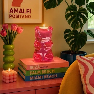 May include: A translucent pink gummy bear-shaped candle with a lit wick, resting on a stack of three books titled Ibiza Bohemia, Palm Beach, and Miami Beach. The candle is positioned on a pink surface next to a small pink bubble candle.