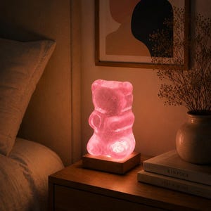 May include: A translucent pink gummy bear-shaped lamp emitting a soft glow, mounted on a square wooden base. The lamp features a smooth, rounded surface with a textured interior, creating a warm light effect that illuminates the surrounding area.
