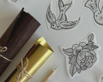 Puede incluir: Una colección de pegatinas de arte lineal en blanco y negro con motivos florales y de aves, un rollo de fieltro marrón oscuro, un rollo de lámina dorada metálica atado con cordel, un punzón de madera y un pequeño rollo de cinta adhesiva estampada en tonos rosa y naranja.