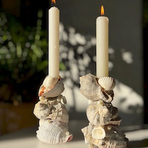 Puede incluir: Dos velas cónicas de color blanco roto se mantienen erguidas mediante bases personalizadas construidas con conchas marinas texturizadas apiladas en tonos beige, tostado y blanco. Pequeñas perlas artificiales color crema se intercalan.