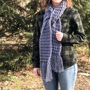 May include: A grey crocheted scarf with fringe. The scarf is worn by a person wearing a green and black checked shirt and blue jeans.