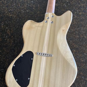 Puede incluir: Vista trasera del cuerpo de una guitarra el&eacute;ctrica con un acabado de madera clara natural. Incluye una tapa negra en forma de l&aacute;grima para la cavidad de control, un puente cromado de cuerdas a trav&eacute;s del cuerpo y un m&aacute;stil atornillado con cuatro tornillos.