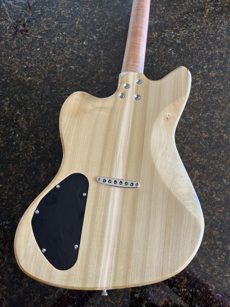 Puede incluir: Vista trasera del cuerpo de una guitarra el&eacute;ctrica con un acabado de madera clara natural. Incluye una tapa negra en forma de l&aacute;grima para la cavidad de control, un puente cromado de cuerdas a trav&eacute;s del cuerpo y un m&aacute;stil atornillado con cuatro tornillos.