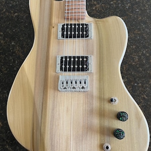 Puede incluir: Una guitarra el&eacute;ctrica con cuerpo de madera clara de veta natural, que presenta dos pastillas humbucker negras, un puente plateado, un interruptor de palanca de tres posiciones, dos perillas de control negras texturizadas con detalles verdes.