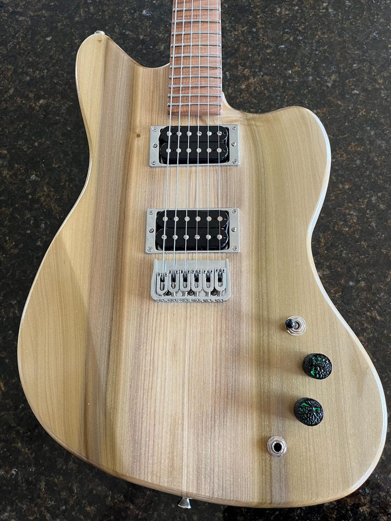 Puede incluir: Una guitarra el&eacute;ctrica con cuerpo de madera clara de veta natural, que presenta dos pastillas humbucker negras, un puente plateado, un interruptor de palanca de tres posiciones, dos perillas de control negras texturizadas con detalles verdes.
