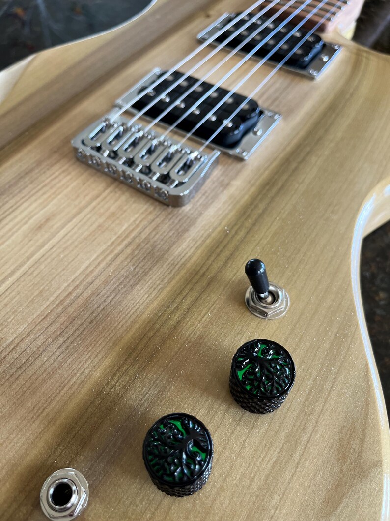 Puede incluir: Un cuerpo de guitarra el&eacute;ctrica de madera clara con veta visible. Cuenta con dos pastillas humbucker negras, un puente plateado, un interruptor de palanca negro y dos perillas de control negras con un dise&ntilde;o de &aacute;rbol de la vida verde.