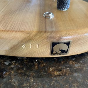 Puede incluir: Un cuerpo de guitarra el&eacute;ctrica de madera clara con un puente, dos perillas de control negras texturizadas y un jack de entrada de metal. El n&uacute;mero 371 est&aacute; estampado en la madera junto a una pegatina cuadrada que muestra un oso polar y MADE IN ALASKA.