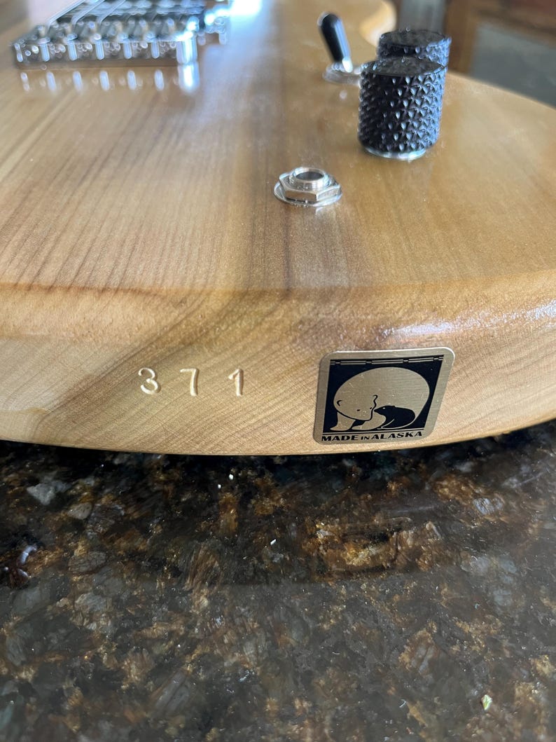 Puede incluir: Un cuerpo de guitarra el&eacute;ctrica de madera clara con un puente, dos perillas de control negras texturizadas y un jack de entrada de metal. El n&uacute;mero 371 est&aacute; estampado en la madera junto a una pegatina cuadrada que muestra un oso polar y MADE IN ALASKA.
