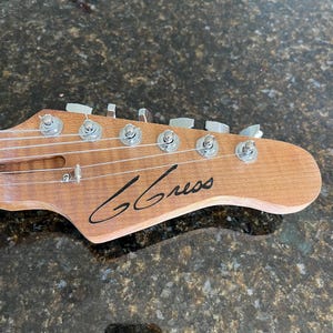 Puede incluir: Un clavijero de guitarra de madera marr&oacute;n claro con seis clavijas de afinaci&oacute;n plateadas, un retenedor de cuerdas y el texto G Gress en cursiva negra impreso en el frente. El clavijero tiene un acabado de veta natural y seis cuerdas.