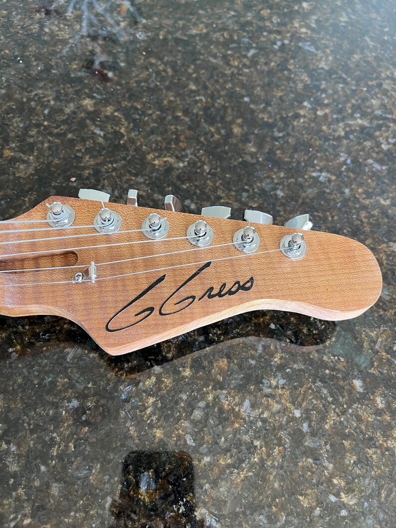 Puede incluir: Un clavijero de guitarra de madera marr&oacute;n claro con seis clavijas de afinaci&oacute;n plateadas, un retenedor de cuerdas y el texto G Gress en cursiva negra impreso en el frente. El clavijero tiene un acabado de veta natural y seis cuerdas.