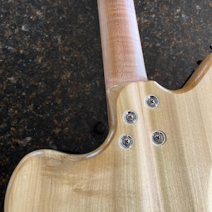 Puede incluir: La parte posterior del cuerpo y la uni&oacute;n del m&aacute;stil de una guitarra el&eacute;ctrica de madera clara. El m&aacute;stil est&aacute; unido al cuerpo con cuatro tornillos met&aacute;licos plateados insertados en casquillos circulares empotrados. La veta es visible.