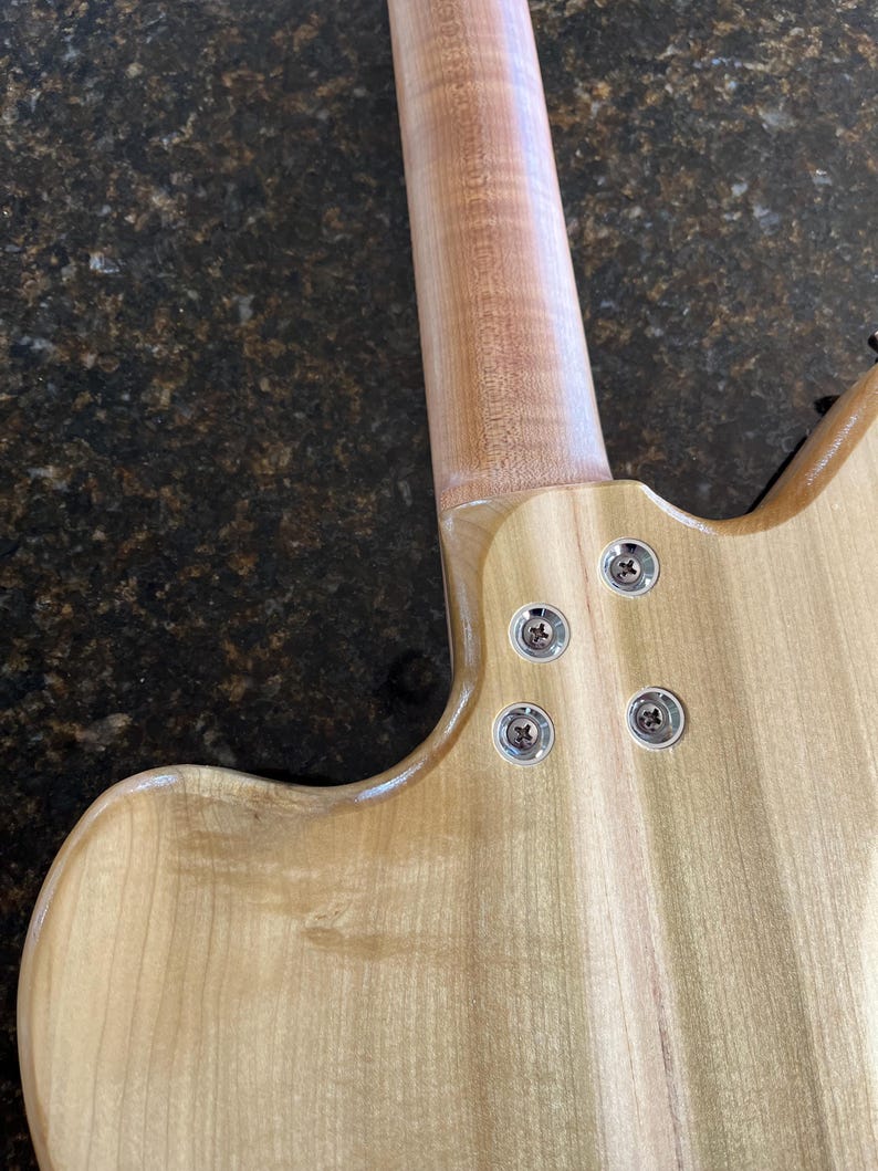 Puede incluir: La parte posterior del cuerpo y la uni&oacute;n del m&aacute;stil de una guitarra el&eacute;ctrica de madera clara. El m&aacute;stil est&aacute; unido al cuerpo con cuatro tornillos met&aacute;licos plateados insertados en casquillos circulares empotrados. La veta es visible.