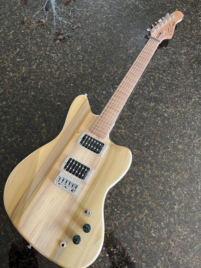 Puede incluir: Una guitarra el&eacute;ctrica con cuerpo de madera clara de veta natural, m&aacute;stil y diapas&oacute;n de arce. Cuenta con dos pastillas humbucker negras, un puente plateado, tres perillas de control y un clavijero con la marca Glarry impresa en negro.