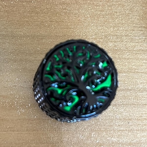 Puede incluir: Un anillo circular de metal negro con un dise&ntilde;o recortado de un &aacute;rbol con ramas y ra&iacute;ces extendidas. El fondo detr&aacute;s de la silueta del &aacute;rbol est&aacute; relleno con un color verde ne&oacute;n vibrante y brillante, creando un efecto visual de alto contraste.