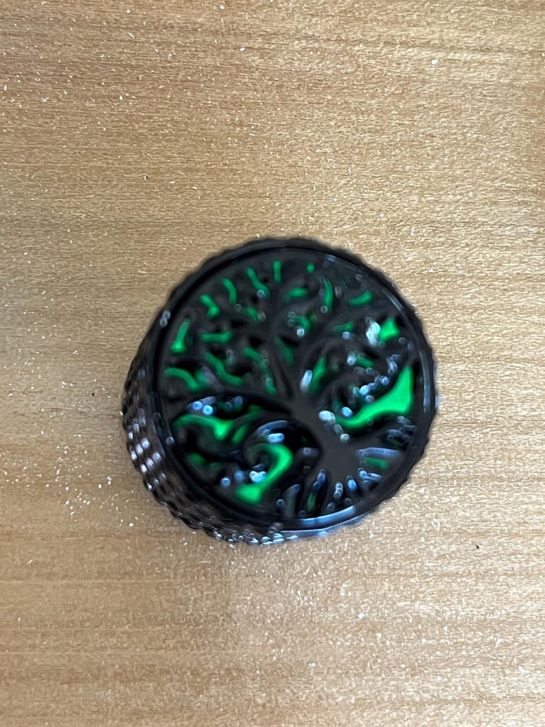Puede incluir: Un anillo circular de metal negro con un dise&ntilde;o recortado de un &aacute;rbol con ramas y ra&iacute;ces extendidas. El fondo detr&aacute;s de la silueta del &aacute;rbol est&aacute; relleno con un color verde ne&oacute;n vibrante y brillante, creando un efecto visual de alto contraste.