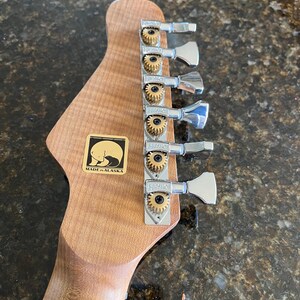 Puede incluir: Un clavijero de guitarra de madera marr&oacute;n claro con seis clavijas de afinaci&oacute;n plateadas Hipshot con engranajes dorados. Una pegatina cuadrada dorada es visible en la madera, mostrando un gr&aacute;fico de silueta de oso negro y el texto MADE IN ALASKA.
