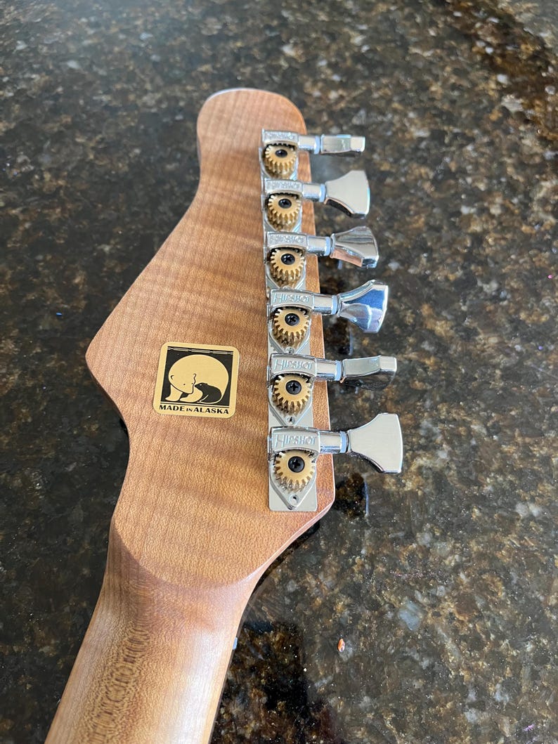 Puede incluir: Un clavijero de guitarra de madera marr&oacute;n claro con seis clavijas de afinaci&oacute;n plateadas Hipshot con engranajes dorados. Una pegatina cuadrada dorada es visible en la madera, mostrando un gr&aacute;fico de silueta de oso negro y el texto MADE IN ALASKA.