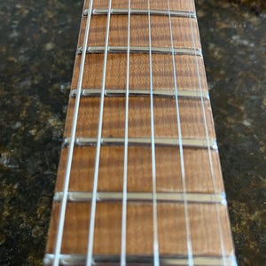 Puede incluir: Un m&aacute;stil de guitarra con un diapas&oacute;n de madera clara flameada con patrones de veta visibles. El m&aacute;stil incluye trastes de metal plateado pulido y seis cuerdas de acero finas y paralelas que corren verticalmente a lo largo del diapas&oacute;n.