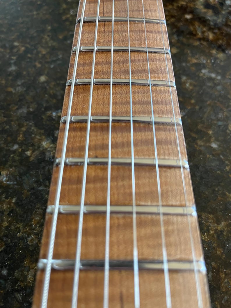 Puede incluir: Un m&aacute;stil de guitarra con un diapas&oacute;n de madera clara flameada con patrones de veta visibles. El m&aacute;stil incluye trastes de metal plateado pulido y seis cuerdas de acero finas y paralelas que corren verticalmente a lo largo del diapas&oacute;n.