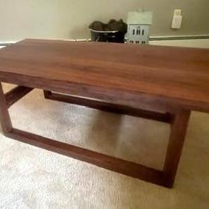 May include: A rectangular coffee table constructed from dark brown wood. The table features a smooth, flat top surface and a base composed of four vertical legs connected by horizontal support bars, creating an open, geometric frame design.