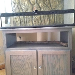 May include: A gray wooden cabinet with two doors and a central open shelf containing a single wooden branch. A large glass terrarium with a black frame and a textured rock-like background sits on top of the cabinet, featuring a central locking mechanism.