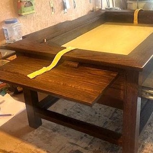 May include: A dark wood table with a pull-out drawer and a large rectangular top. The table has a light brown surface and a dark brown frame. The table is in a workshop setting.