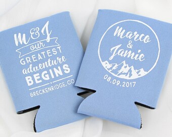 May include: Two blue and white can coolers with a mountain theme. One cooler says "M&J our greatest adventure begins Breckenridge, CO". The other cooler says "Marco & Jamie 08.09.2017" with a circle around the names and mountains inside the circle.