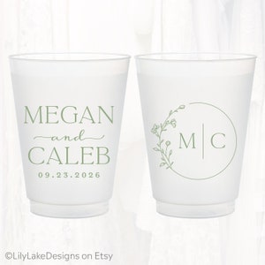 Pu&ograve; includere: Due bicchieri in plastica satinata con testo e grafiche color verde salvia. Il bicchiere a sinistra riporta i nomi Megan e Caleb con la data 23.09.2026. Il bicchiere a destra mostra una corona floreale circolare che racchiude le iniziali M e C.
