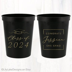 May include: Two black plastic cups with gold lettering. One cup says "Class of 2024" with a graduation cap icon. The other cup says "Congrats Jessica SHS Grad" in a rectangular frame.