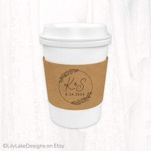 May include: A white paper coffee cup with a brown sleeve featuring a black floral wreath design. The wreath encircles the initials "K & S" and the date "8.24.2024". The cup has a white lid.