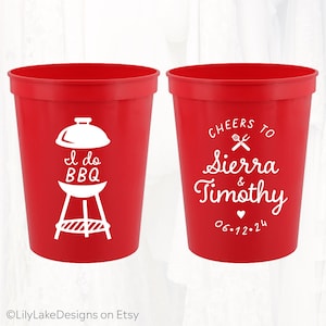 May include: Two red plastic cups with white text and graphics. One cup says "I do BBQ" with a grill graphic. The other cup says "Cheers to Sierra & Timothy 06.12.24" with a fork and knife graphic.