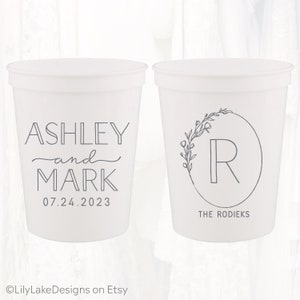 May include: Two white plastic cups with black text. One cup says "Ashley and Mark 07.24.2023". The other cup has a floral wreath with the letter "R" inside and the text "The Rodieks".