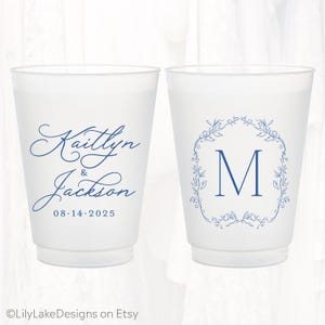 May include: Two frosted plastic cups featuring blue ink designs. The left cup displays the names Kaitlyn & Jackson with the date 08.14.2025 in script and serif fonts. The right cup features a large letter M inside a blue floral wreath design.