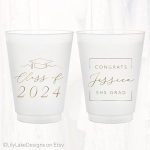 May include: Two white plastic cups with gold lettering. One cup says "Class of 2024" with a graduation cap graphic. The other cup says "Congrats Jessica SHS GRAD" inside a gold frame. Ideal for graduation parties.