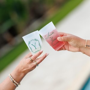 May include: Two frosted plastic cups held up for a toast. One cup features a green botanical wreath surrounding the letter Y. The other cup contains a pink liquid and displays the text Sara & Joe 06-26-2026 in a dark green cursive script.