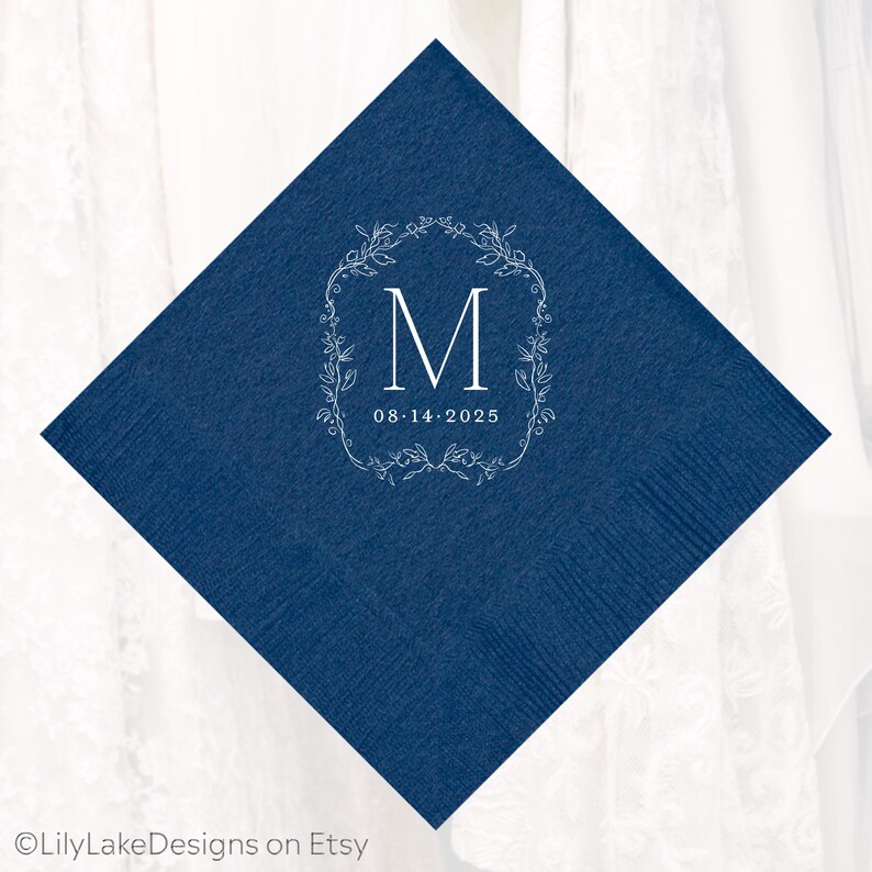 Puede incluir: Una servilleta cuadrada azul marino con un dise&ntilde;o de corona floral blanca. La corona rodea la letra "M" y la fecha "08.14.2025". La servilleta est&aacute; hecha de un material texturizado, ideal para una boda o evento especial.