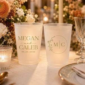 Pu&ograve; includere: Due bicchieri in plastica satinata con testo verde personalizzato. Un bicchiere mostra i nomi Megan e Caleb con la data 23.09.2026. L'altro presenta un design a corona botanica circolare che circonda le iniziali M e C separate da una linea.