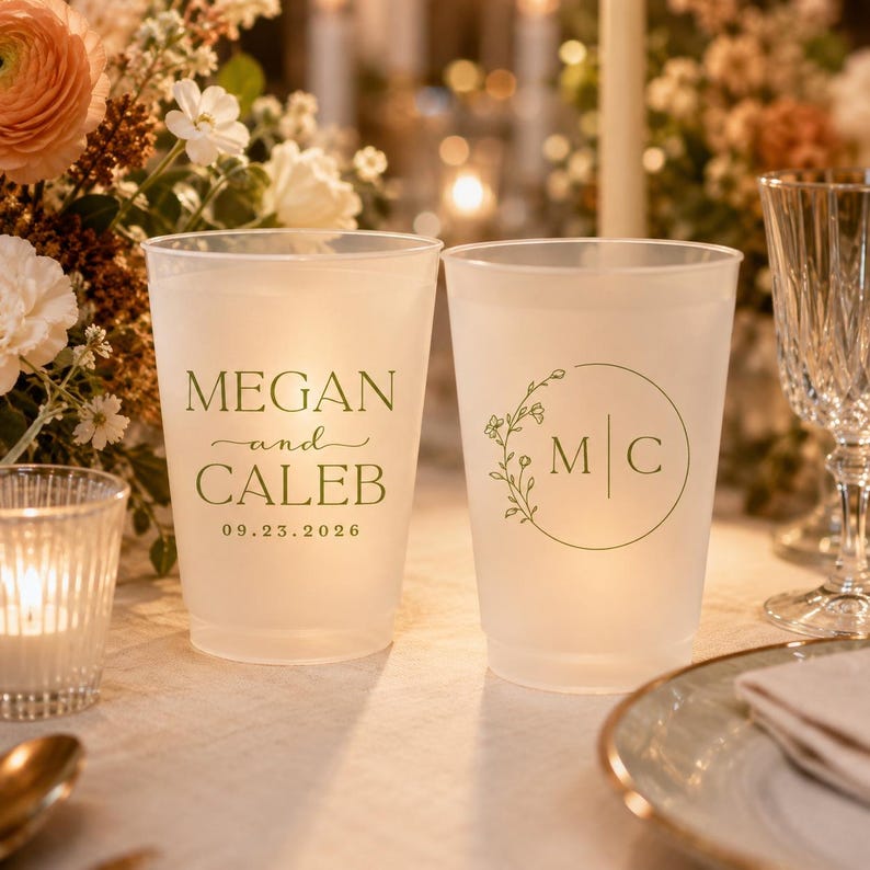 Pu&ograve; includere: Due bicchieri in plastica satinata con testo verde personalizzato. Un bicchiere mostra i nomi Megan e Caleb con la data 23.09.2026. L'altro presenta un design a corona botanica circolare che circonda le iniziali M e C separate da una linea.