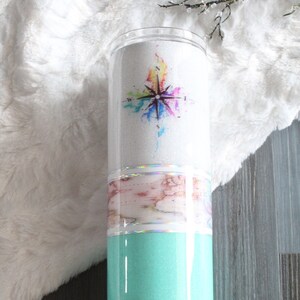 Puede incluir: Un vaso turquesa con un diseño de purpurina blanca y dorada. El vaso presenta una brújula de acuarela con un diseño de arcoíris y un mapa del mundo.