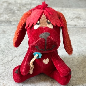May include: A vintage red plush dog toy with orange ears and a red head covering. The dog has a black nose, a blue flower, and a white heart. The toy is sitting on a gray surface.