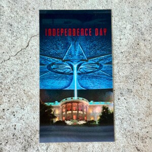 May include: A rectangular postcard featuring a scene from the movie "Independence Day." The top half shows a blue alien spacecraft above the White House, which is lit up at night. The title "INDEPENDENCE DAY" is in red letters.